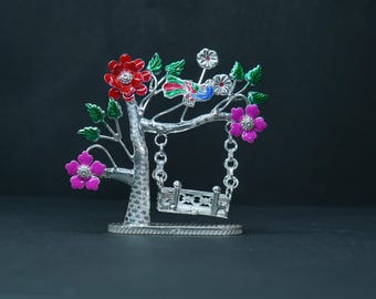 May include: A decorative silver-colored tree sculpture with a swing. The tree has red, pink, and white flowers, green leaves, and a colorful bird. The swing is suspended from the branches by chains.