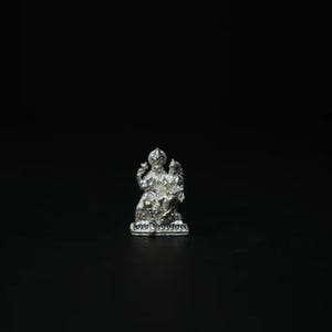 May include: A small, detailed silver statue of a deity, possibly Hindu, against a black background. The statue depicts a seated figure with multiple arms and ornate headwear, showcasing intricate craftsmanship.
