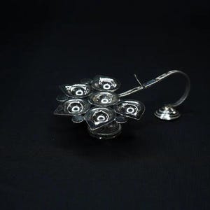 May include: A silver-colored, multi-cup oil lamp with a curved handle and a small, round base. The lamp has a flower-like design with several small, shallow cups for holding oil. The background is a solid black.