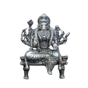 May include: A silver-colored statue of a deity seated on a decorative bench. The statue has multiple arms and is adorned with intricate jewelry and a detailed headdress. The figure is set against a plain white background.