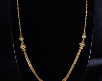May include: A gold-colored necklace with a delicate chain design. The necklace features decorative cone-shaped elements on each side, connected to a thicker chain that drapes in a gentle curve. The necklace is displayed against a black background.