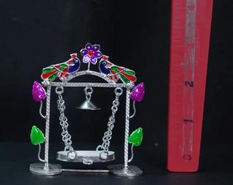 May include: A silver-colored decorative swing set with two colorful peacocks perched on top. The swing has a small bell hanging from the center and is adorned with purple, green, and red accents. The swing is approximately 2 inches tall.