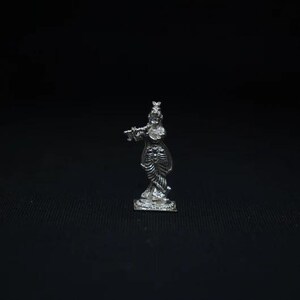 Puede incluir: Pequeña figura plateada de una persona tocando la flauta. La figura está sobre una base rectangular, con detalles intrincados en la ropa y el cabello. El fondo es negro liso.