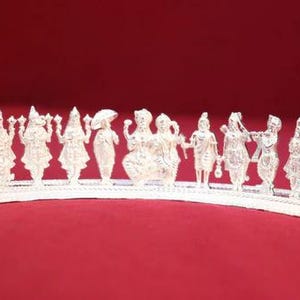 May include: A silver-colored, decorative piece featuring multiple figurines of deities and figures, arranged in a row. The figures are intricately detailed and stand on a curved base. The background is a solid red color.