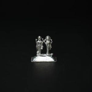 May include: A small, silver-colored statue depicting two figures standing side-by-side on a rectangular base. The figures are detailed with intricate designs and stand against a black background. The statue appears to be a religious or cultural artifact.