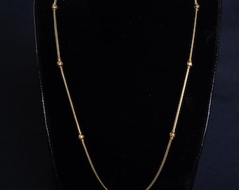 May include: A gold-colored necklace with a delicate chain design. The necklace features small, round beads evenly spaced along the chain. The necklace is displayed on a black mannequin.