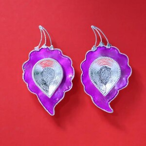 May include: Two decorative leaf-shaped dishes with a vibrant purple hue and silver accents. Each dish features a silver, teardrop-shaped interior with intricate patterns. The dishes have a silver loop at the top and are set against a red background.