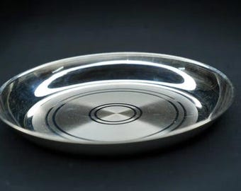 May include: A shiny, oval-shaped stainless steel plate. The plate has a polished surface with concentric circles and a central design. The plate is set against a dark background, highlighting its reflective qualities.