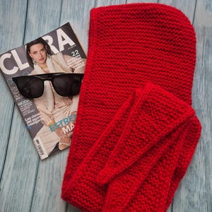 May include: A bright red knitted hood and scarf set. The hood is rectangular with a rounded top, and the scarf has long, pointed ends. A magazine with the word "CLARA" and sunglasses are next to the set.
