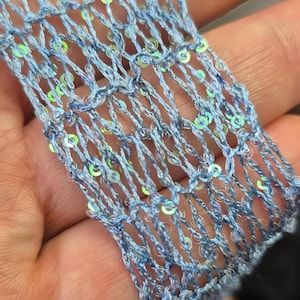 May include: Close-up of a light blue, open-weave fabric with iridescent sequins. The fabric appears to be a decorative trim or embellishment, possibly for clothing or crafts. The sequins add a shimmering effect.