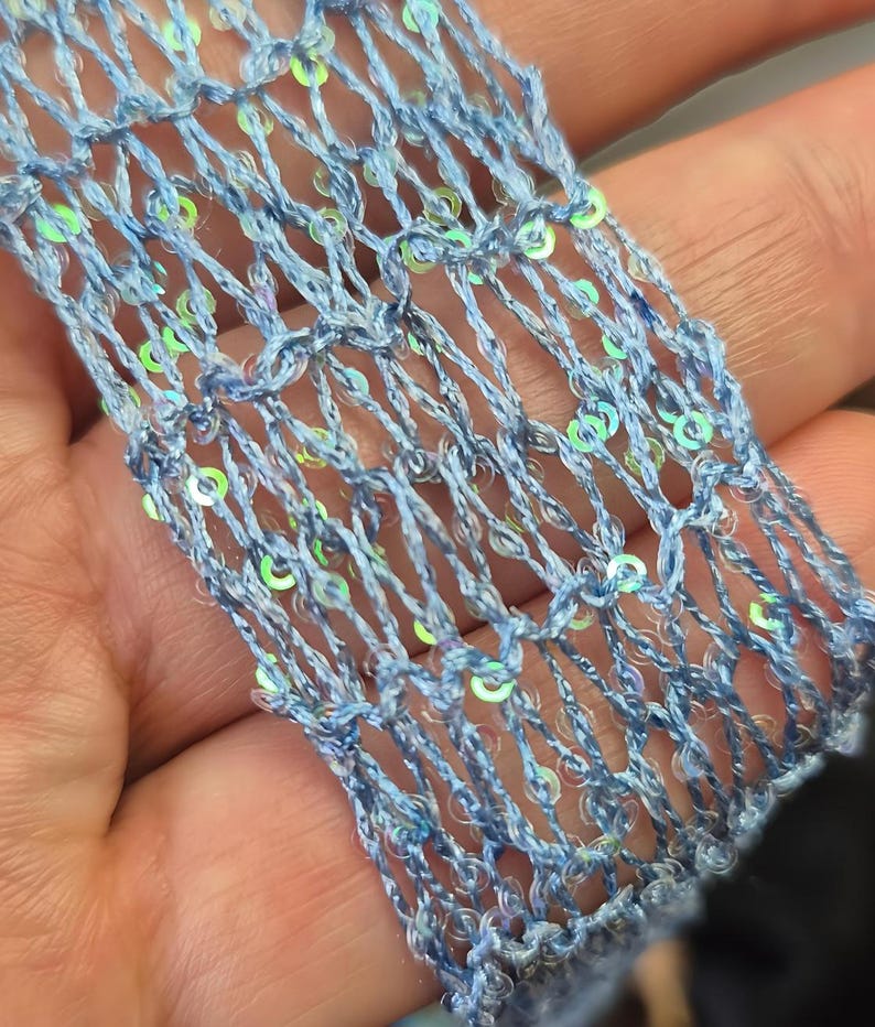 May include: Close-up of a light blue, open-weave fabric with iridescent sequins. The fabric appears to be a decorative trim or embellishment, possibly for clothing or crafts. The sequins add a shimmering effect.