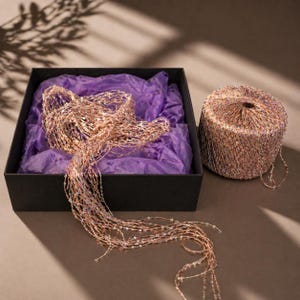 May include: A black box with purple tissue paper holds a shimmering, beaded garland. A cylindrical spool of the same garland sits beside the box. The garland is a mix of gold, copper, and iridescent beads, creating a festive, decorative look.
