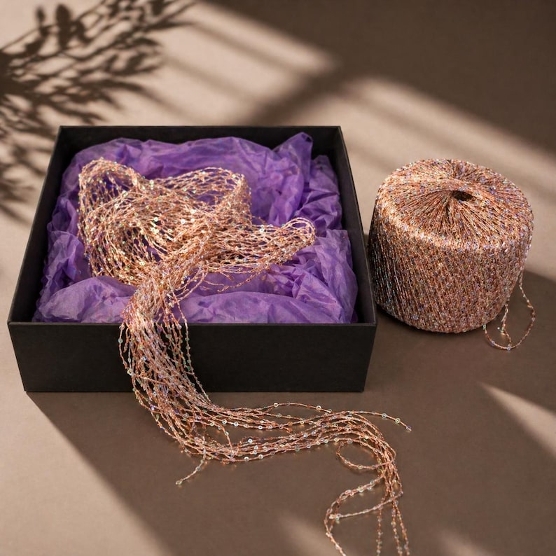May include: A black box with purple tissue paper holds a shimmering, beaded garland. A cylindrical spool of the same garland sits beside the box. The garland is a mix of gold, copper, and iridescent beads, creating a festive, decorative look.