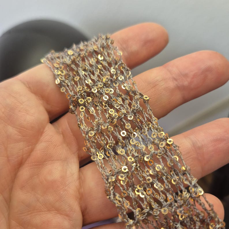 May include: Close-up of a decorative trim featuring a net-like design. The trim is a light gray color with gold sequins. The sequins are evenly spaced throughout the trim, adding a touch of sparkle. This trim is suitable for various craft and sewing projects.