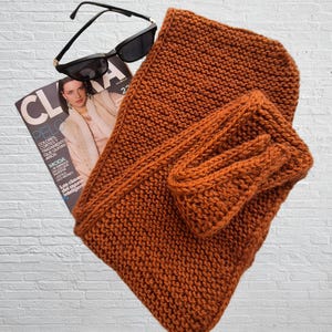 Könnte beinhalten: Ein orangefarbenes Strickschal- und passendes Handgelenkwärmer-Set. Der Schal ist gefaltet und der Handgelenkwärmer liegt obenauf. Eine schwarze Sonnenbrille und eine Zeitschrift mit dem Wort "CLA" sind ebenfalls im Bild.