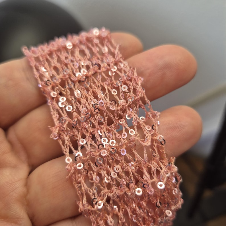 May include: Close-up of a piece of pink, open-weave fabric adorned with small, shimmering sequins. The fabric has a delicate, net-like appearance, with the sequins catching the light. The overall effect is one of subtle sparkle and texture.