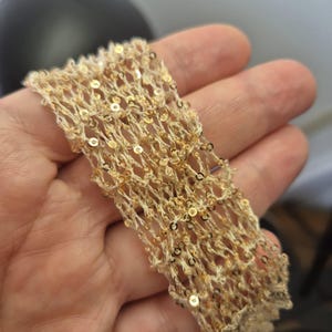 May include: Close-up of a beige, open-weave fabric strip adorned with small, gold sequins. The fabric has a delicate, net-like appearance, with the sequins adding a touch of sparkle. The strip is held in a hand, showcasing its texture and detail.