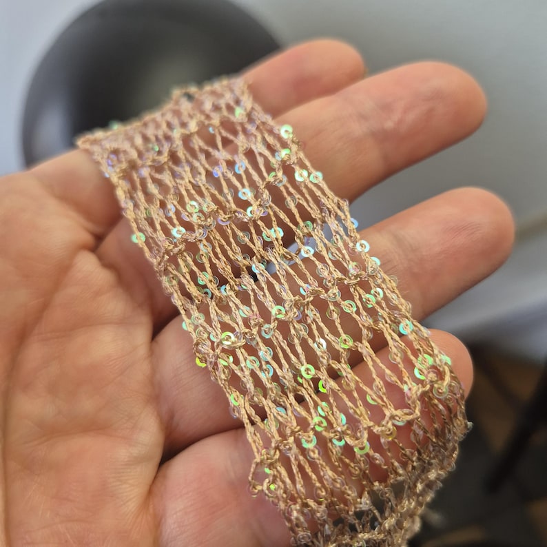 May include: Close-up of a beige, open-weave fabric strip adorned with iridescent sequins. The fabric has a net-like appearance, with the sequins catching the light. The strip is held in a hand, showcasing the texture and detail of the material.