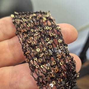 May include: Close-up of a black mesh fabric adorned with gold sequins. The fabric is held in a hand, showcasing the intricate weave and the shimmering effect of the sequins. The texture appears delicate and potentially suitable for embellishments.