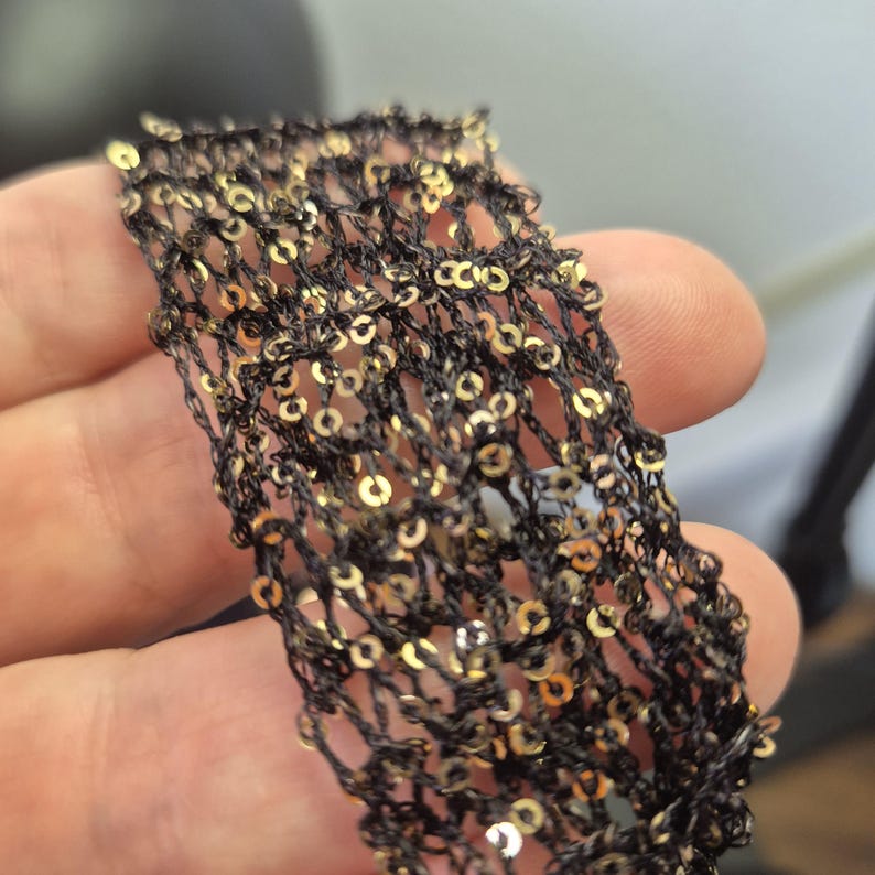 May include: Close-up of a black mesh fabric adorned with gold sequins. The fabric is held in a hand, showcasing the intricate weave and the shimmering effect of the sequins. The texture appears delicate and potentially suitable for embellishments.