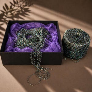 May include: A black box with a necklace made of small, iridescent sequins. The necklace is draped over purple tissue paper. A spool of matching sequin material sits next to the box. The sequins reflect shades of green, blue, and silver.