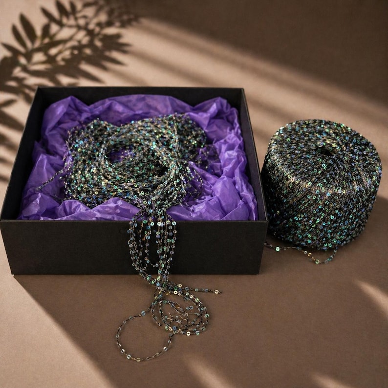 May include: A black box with a necklace made of small, iridescent sequins. The necklace is draped over purple tissue paper. A spool of matching sequin material sits next to the box. The sequins reflect shades of green, blue, and silver.