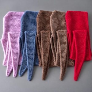 Könnte beinhalten: Fünf gestrickte Schals, die nebeneinander in einer Reihe angeordnet sind. Die Schals sind einfarbig und in den Farben Hellrosa, Staubblau, zwei Brauntönen und Hellrot gehalten. Jeder Schal hat eine markante kraus rechts gestrickte Textur und lange, spitz zulaufende Enden.