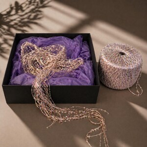 May include: A black box with purple tissue paper holds a beaded garland. A spool of matching yarn sits beside the box. The garland and yarn are a mix of gold, pink, and iridescent beads. The scene is lit by natural light.