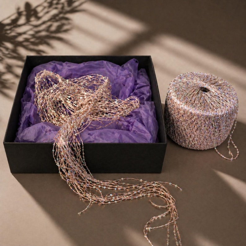 May include: A black box with purple tissue paper holds a beaded garland. A spool of matching yarn sits beside the box. The garland and yarn are a mix of gold, pink, and iridescent beads. The scene is lit by natural light.