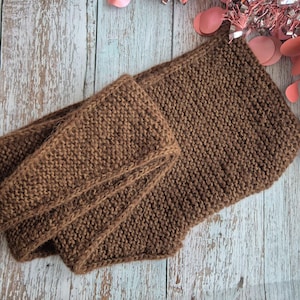 May include: A brown knitted scarf and cowl set. The scarf is long and rectangular, while the cowl is a shorter, wider loop. Both pieces are made of a chunky knit fabric. The set is displayed on a light-colored wooden surface.