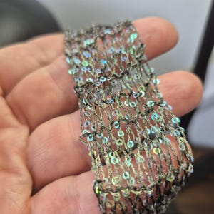 May include: Close-up of a section of decorative trim. The trim is a net-like design with iridescent sequins. The base color of the trim is a dark gray, and the sequins reflect a rainbow of colors. The trim is held in a hand.