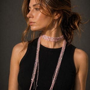 May include: A woman wearing a black tank top and a layered pink beaded necklace. The necklace has multiple strands of small beads that cascade down the chest. The background is a dark gray.