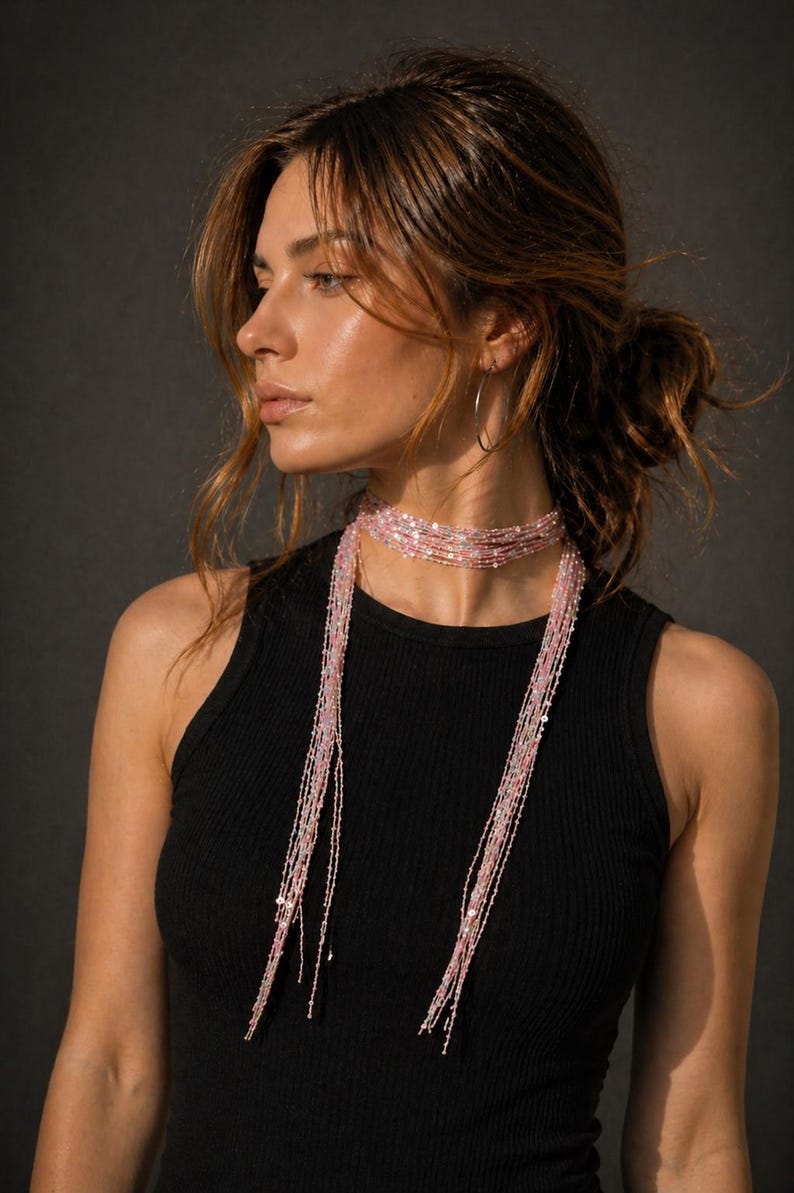 May include: A woman wearing a black tank top and a layered pink beaded necklace. The necklace has multiple strands of small beads that cascade down the chest. The background is a dark gray.