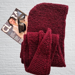 May include: A knitted burgundy scarf and hood set. A fashion magazine and black sunglasses rest on the scarf. The magazine cover features a woman with the word "CLARA" at the top. The scarf is made of thick, textured yarn.