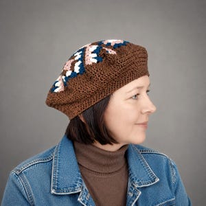 Puede incluir: Una boina de ganchillo marrón con un patrón geométrico en la parte superior, con detalles en hilo blanco, rosa claro y azul oscuro. El gorro está construido con una base marrón sólida y un patrón de punto texturizado e intrincado.