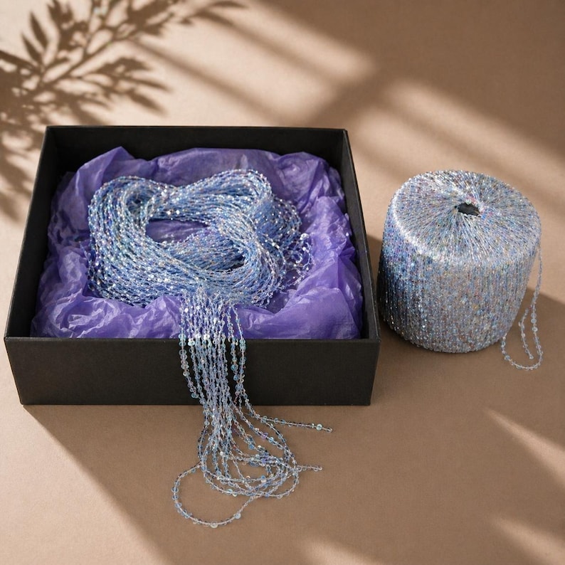 May include: A black box with a bundle of light blue beaded strands and a roll of matching beads. The box contains the strands on purple tissue paper. The roll of beads is cylindrical. The background is a neutral tan color.