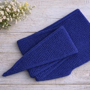 Puede incluir: Un conjunto de bufanda y gorro de punto azul oscuro, exhibido sobre una superficie de madera clara. La bufanda está doblada y el gorro tiene un extremo puntiagudo. Un pequeño ramo de flores blancas y amarillas está en la esquina superior izquierda.