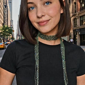 Puede incluir: Una mujer con una camiseta negra y un collar de cuentas verdes. El collar es estilo gargantilla con hebras largas y colgantes. El fondo muestra una calle de la ciudad con edificios altos y un taxi amarillo.