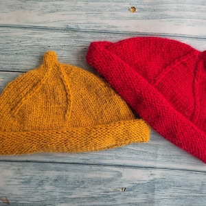 May include: Two knitted hats. One is mustard yellow, the other is bright red. Both hats have a rounded crown and a folded brim. The hats are made of a thick, textured yarn, and are displayed on a light blue wooden surface.