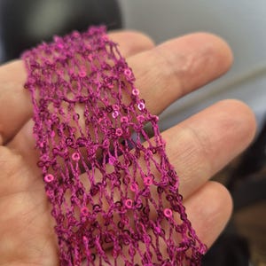 May include: Close-up of a strip of magenta-colored, open-weave fabric. The fabric is embellished with small, round, pink sequins. The texture of the fabric is visible, with a net-like pattern. The sequins add a touch of sparkle.