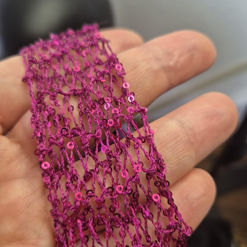 May include: Close-up of a strip of magenta-colored, open-weave fabric. The fabric is embellished with small, round, pink sequins. The texture of the fabric is visible, with a net-like pattern. The sequins add a touch of sparkle.