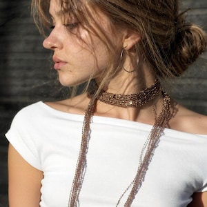 May include: A woman wearing a white off-the-shoulder top and a layered beaded necklace. The necklace is a mix of brown and gold tones, with a choker style and long strands. A silver hoop earring is visible.