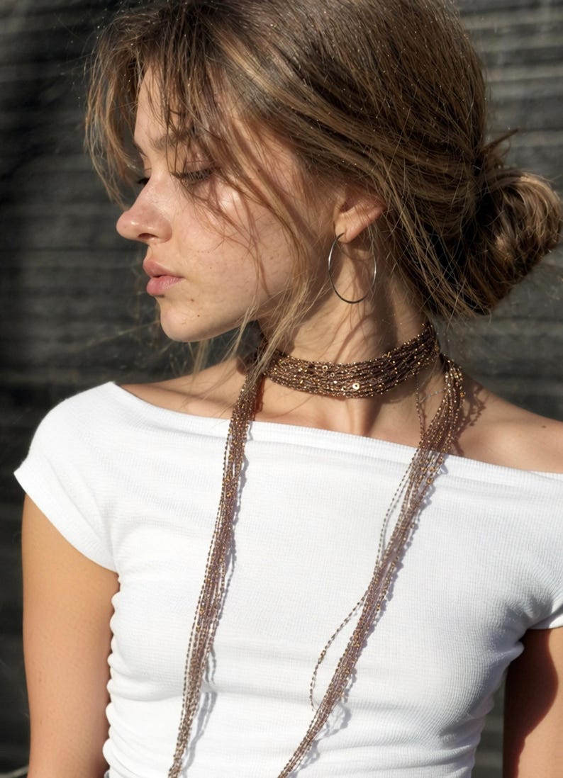 May include: A woman wearing a white off-the-shoulder top and a layered beaded necklace. The necklace is a mix of brown and gold tones, with a choker style and long strands. A silver hoop earring is visible.