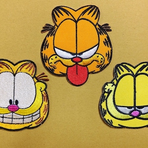 May include: Three embroidered patches featuring the face of a cartoon cat. The top patch shows an orange cat with a red tongue sticking out. The bottom left patch is a yellow cat with a wide grin. The bottom right patch is a yellow cat with eyes closed.