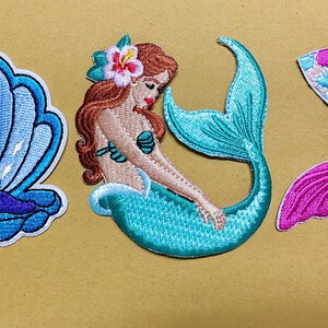 May include: Three embroidered patches featuring a blue seashell with a white pearl, a mermaid with long brown hair and a teal tail adorned with a hibiscus flower, and a mermaid tail with pink and blue scales and a solid pink fin.