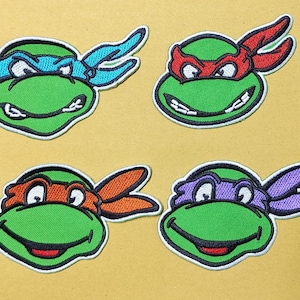 May include: Four embroidered patches featuring the faces of green cartoon turtles. Each turtle wears a different colored eye mask: one blue, one red, one orange, and one purple. The patches have black outlines and white borders.
