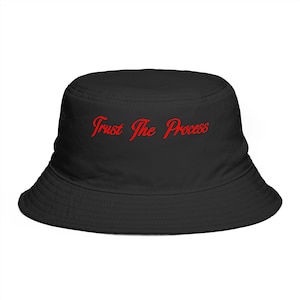 May include: Black bucket hat featuring the red cursive text "Trust The Process". The hat is made of a soft material and has a wide brim. A fashionable accessory for casual wear.