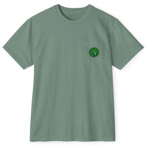 May include: Sage green t-shirt with a crew neck and short sleeves. A small pocket on the chest features a circular graphic with a green and white design. The shirt is made of a soft, comfortable material.