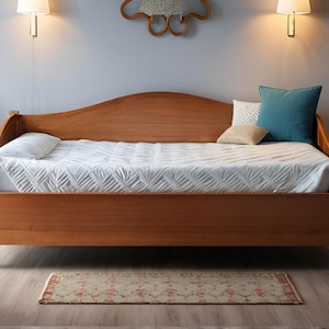 May include: A wooden daybed with a curved headboard and footboard. The bed is made of light brown wood and is covered with a white patterned quilt. Decorative pillows in blue, white, and tan are arranged on the bed. A small rug is on the floor.