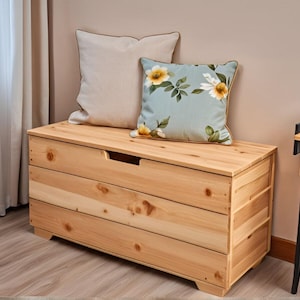 Peut inclure: Un coffre de rangement en bois naturel avec un couvercle à charnières et trois tiroirs. Le coffre est décoré de deux coussins, un blanc et un autre à motif floral.
